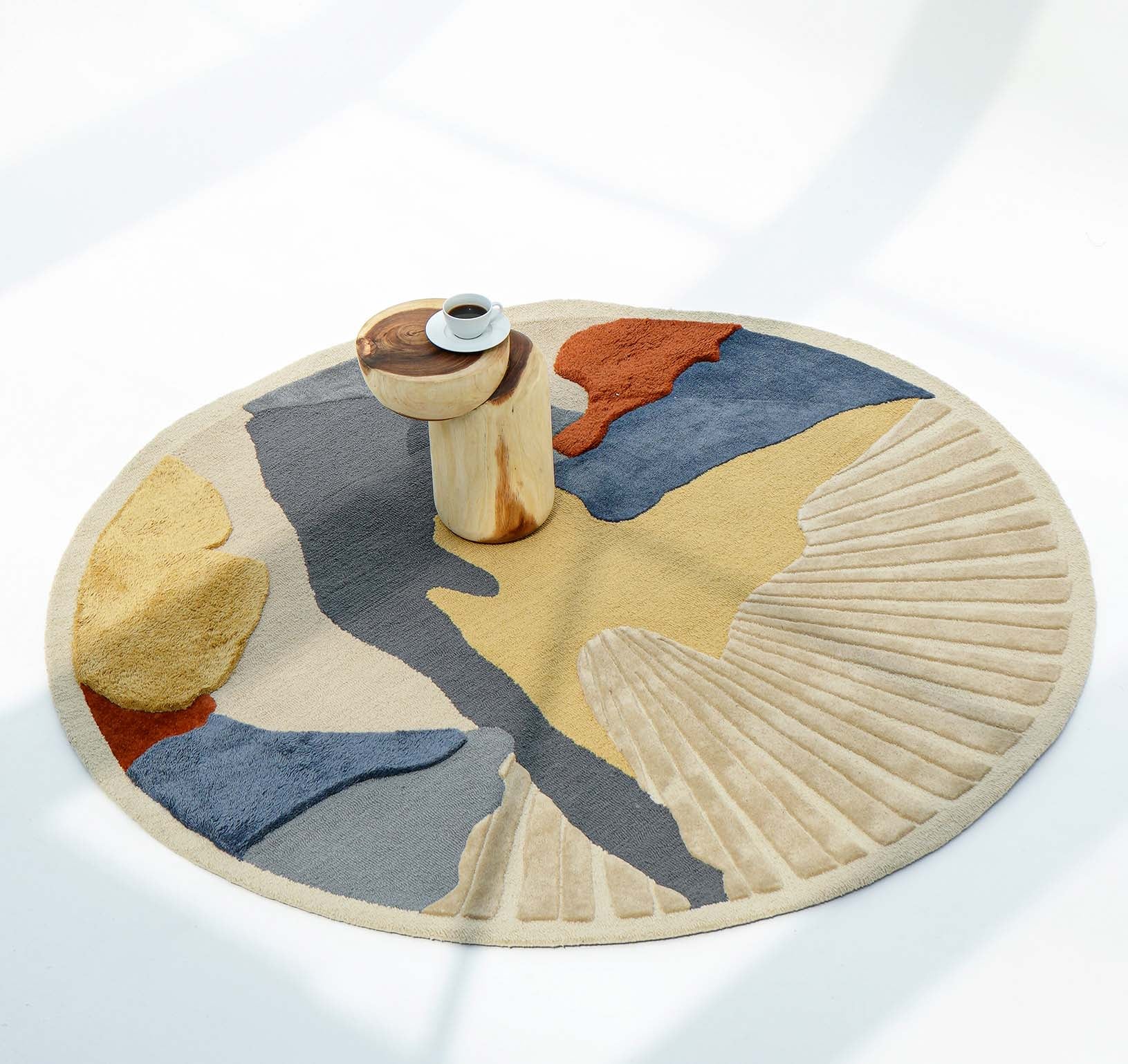 The RAMSHA HOME Multicolor Handmade Round Rug HTW 59 (200cm) features abstract shapes in beige, blue, yellow, and rust. A small wooden side table with a cup and saucer sits atop the rug, set against a bright white background.