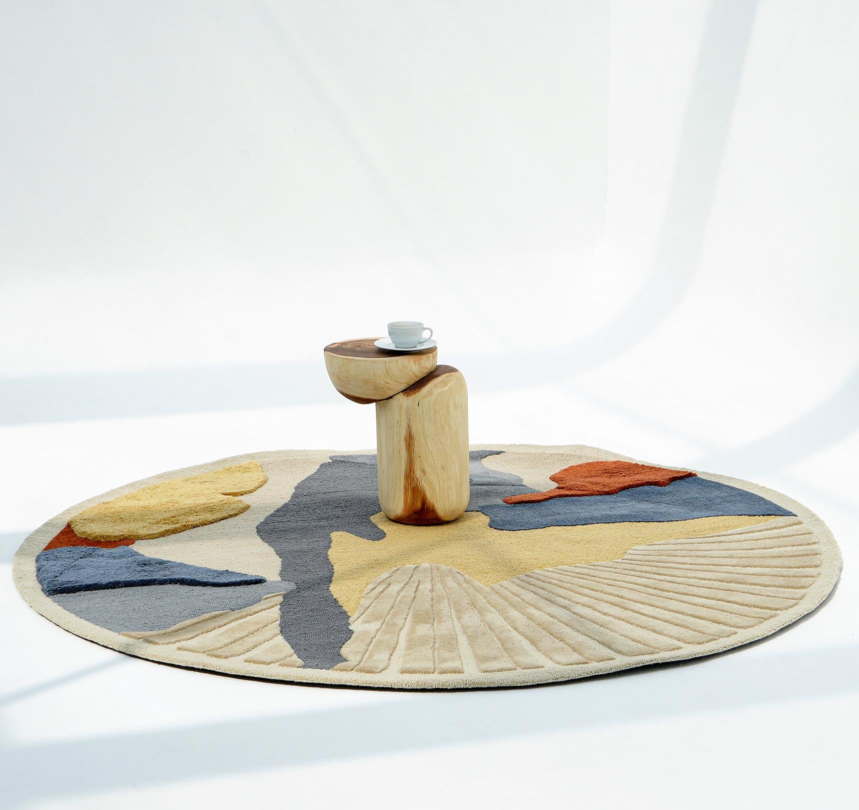 The Multicolor Handmade Round Rug HTW 59 (200cm) by RAMSHA HOME lies beneath a sculptural wooden side table with a white cup and saucer, set against a bright, minimalist backdrop.