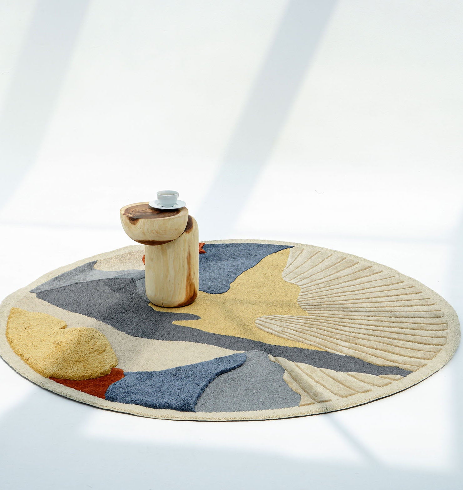 The RAMSHA HOME Multicolor Handmade Round Rug HTW 59 (200cm) features a modern abstract pattern in yellow, blue, gray, and beige, adding style beneath a wooden side table. Sunlight casts soft shadows across the hand-tufted wool rug.