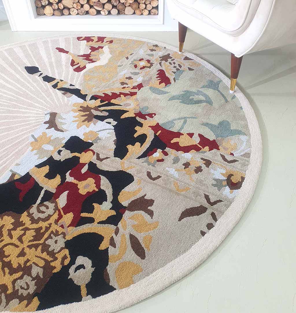 A close-up of the RAMSHA HOME Round multi area rug 200 cm, model HTW-73, features an abstract floral pattern in beige, black, red, gold, and blue tones on a light floor beside a white chair with wooden legs. Firewood is stacked in the background.