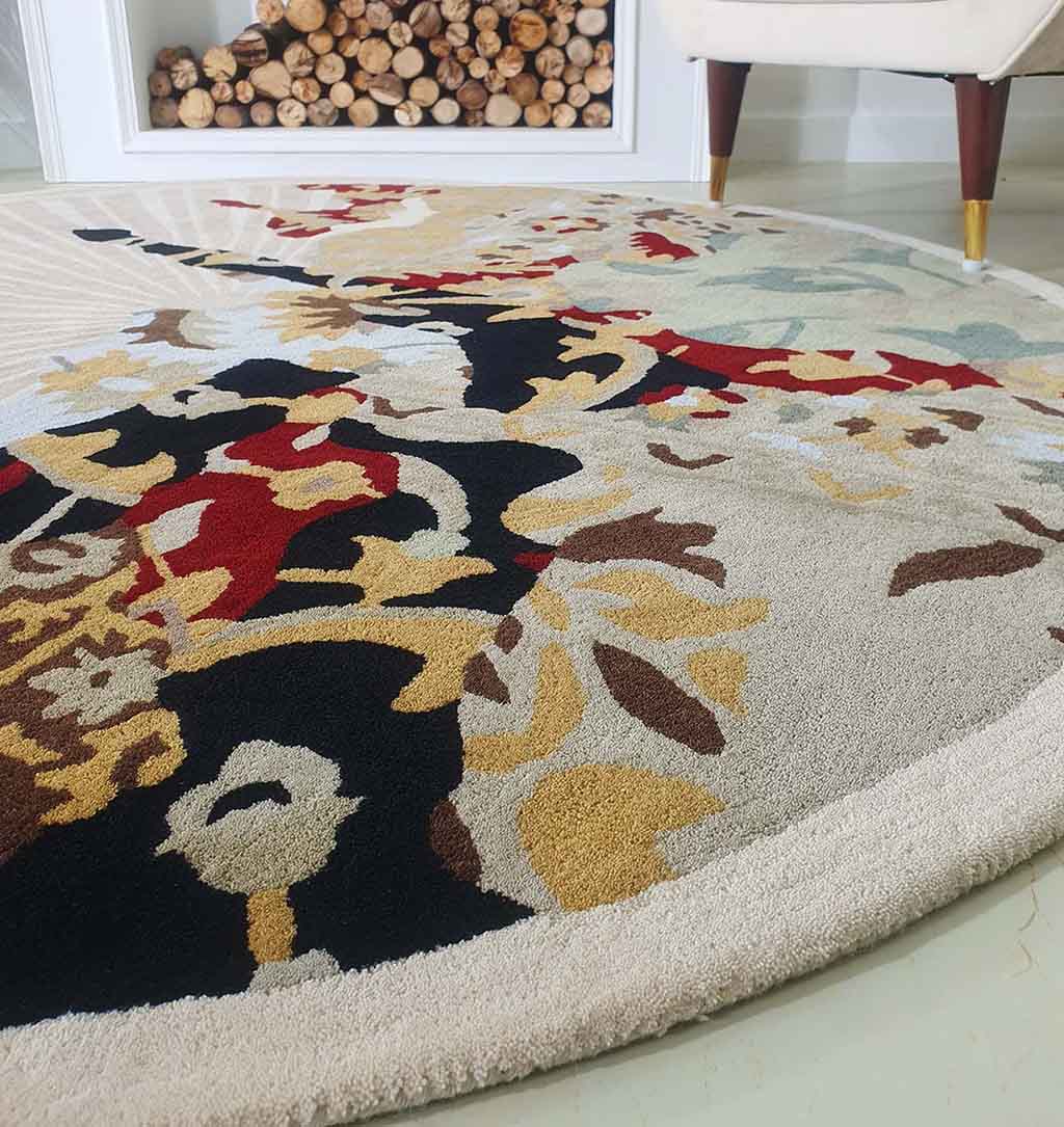 A close-up of the RAMSHA HOME Round Multi Area Rug 200 cm (HTW-73), hand-tufted with beige, black, red, and gold floral designs. A white fireplace with stacked firewood and part of a cream chair with wooden legs are visible behind it.