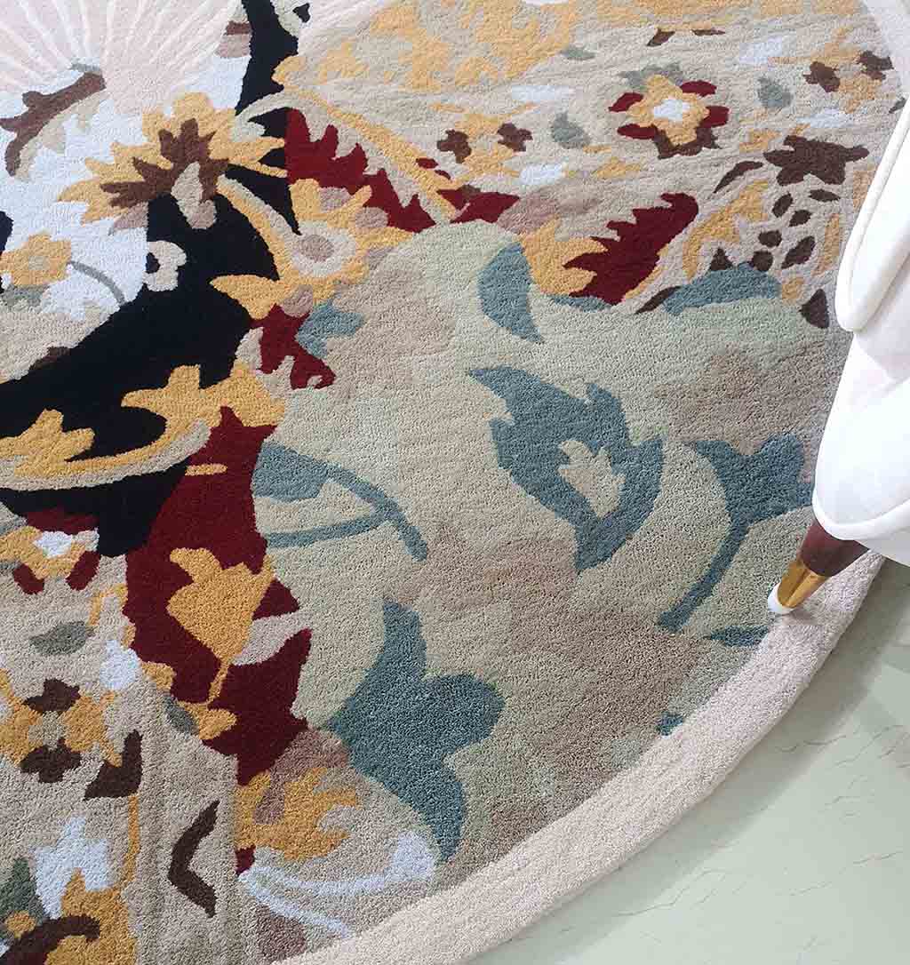 A close-up of the RAMSHA HOME Round multi area rug 200 cm (HTW-73), hand-tufted with an abstract floral pattern in beige, blue, black, yellow, and burgundy, partially covered by a chair leg draped with white fabric.