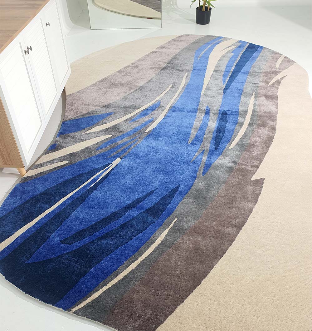 The Irregular Shape Hand-Tufted Carpet HTWV-69 by RAMSHA HOME, featuring abstract blue, gray, and beige patterns, lies on a light floor near a wooden cabinet and potted plant, with a mirror in the background.