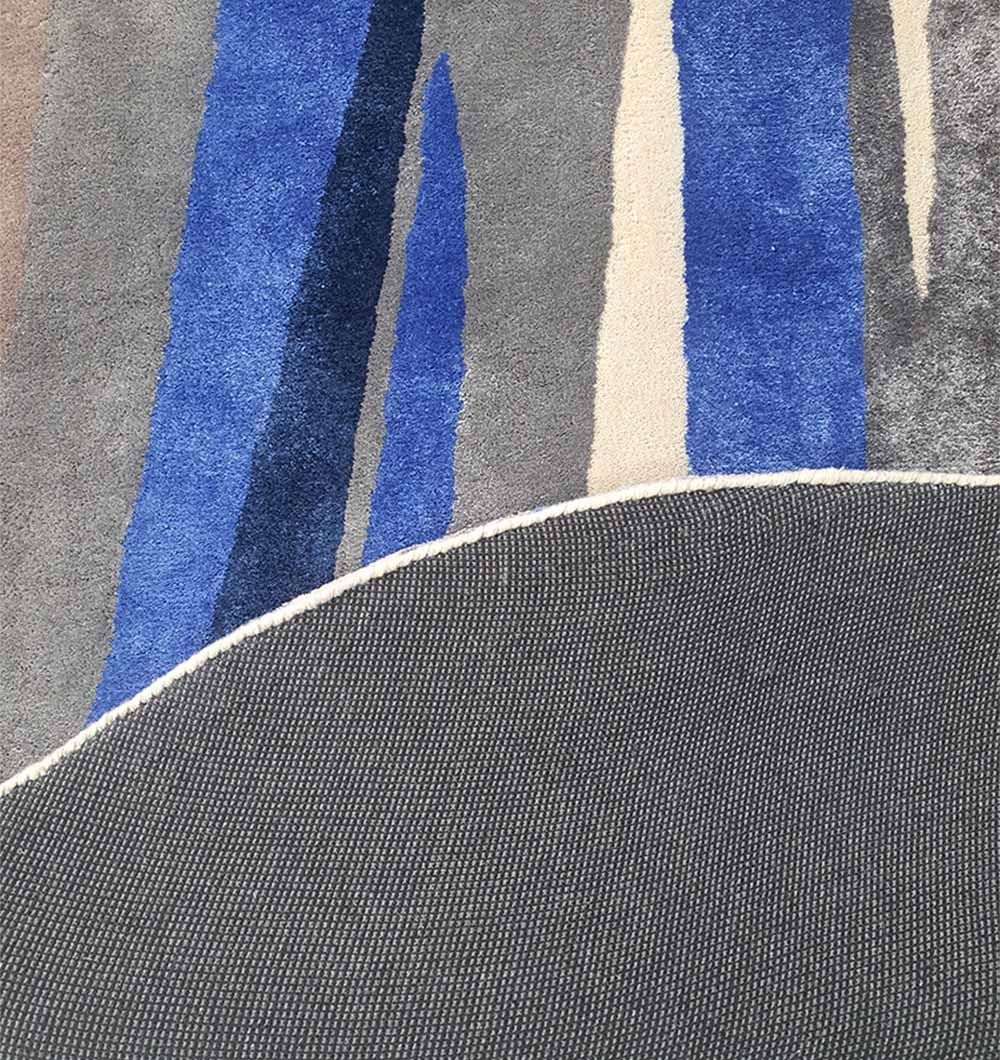 A close-up of the RAMSHA HOME Irregular Shape Hand-Tufted Carpet HTWV-69 shows its abstract blue, gray, and cream patterns with a curved edge folded to reveal the textured dark gray backing, highlighting its quality craftsmanship.
