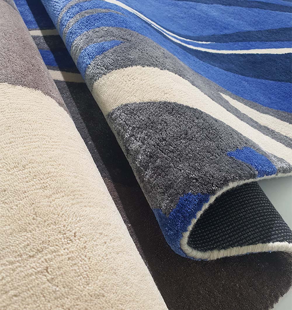 A close-up of two overlapping RAMSHA HOME rugs, including the Irregular Shape Hand-Tufted Carpet HTWV-69 with abstract blue, cream, black, and gray designs. One thick, textured rug is partially folded to show its underside.