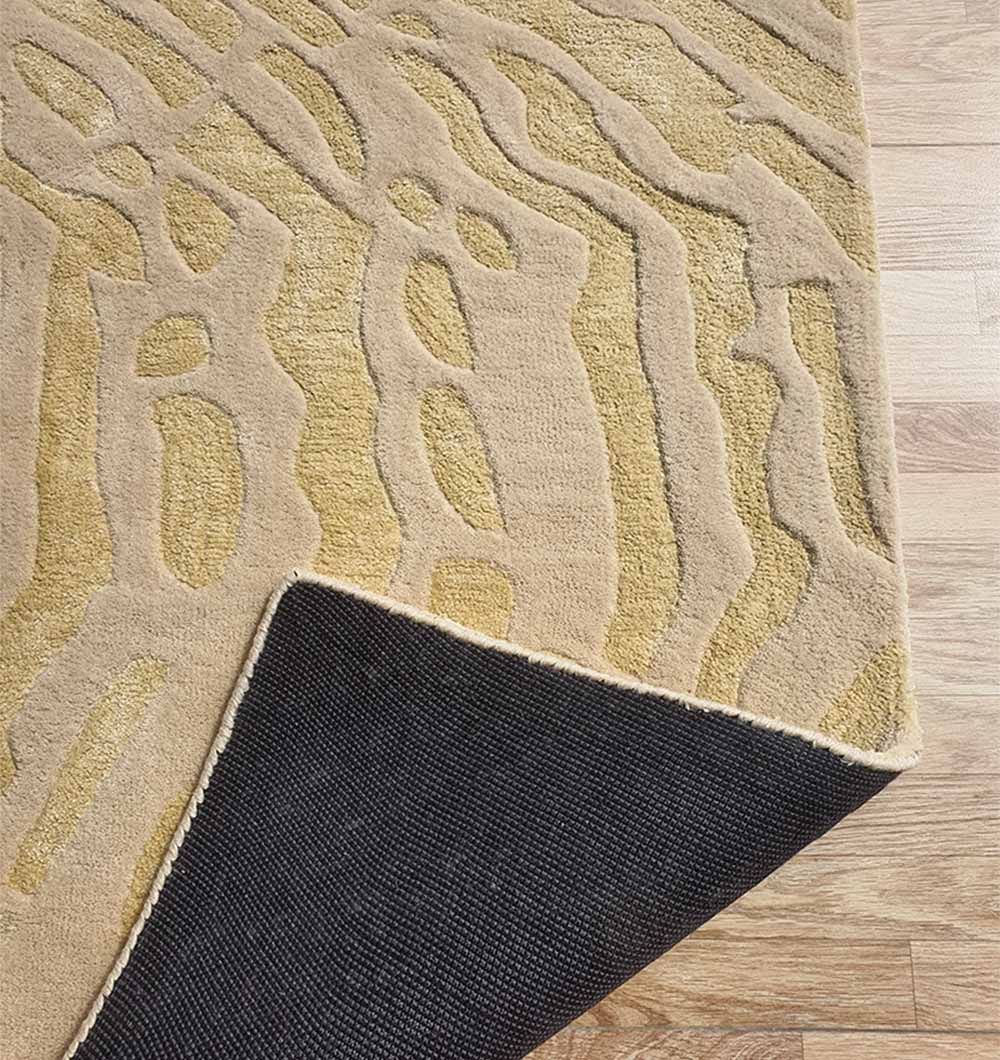 The RAMSHA HOME Handmade Multi Color Carpet For Home Decor (HTWV-75) is displayed on a wooden floor, featuring an abstract beige and gold pattern. One corner is flipped to show its black non-slip backing.