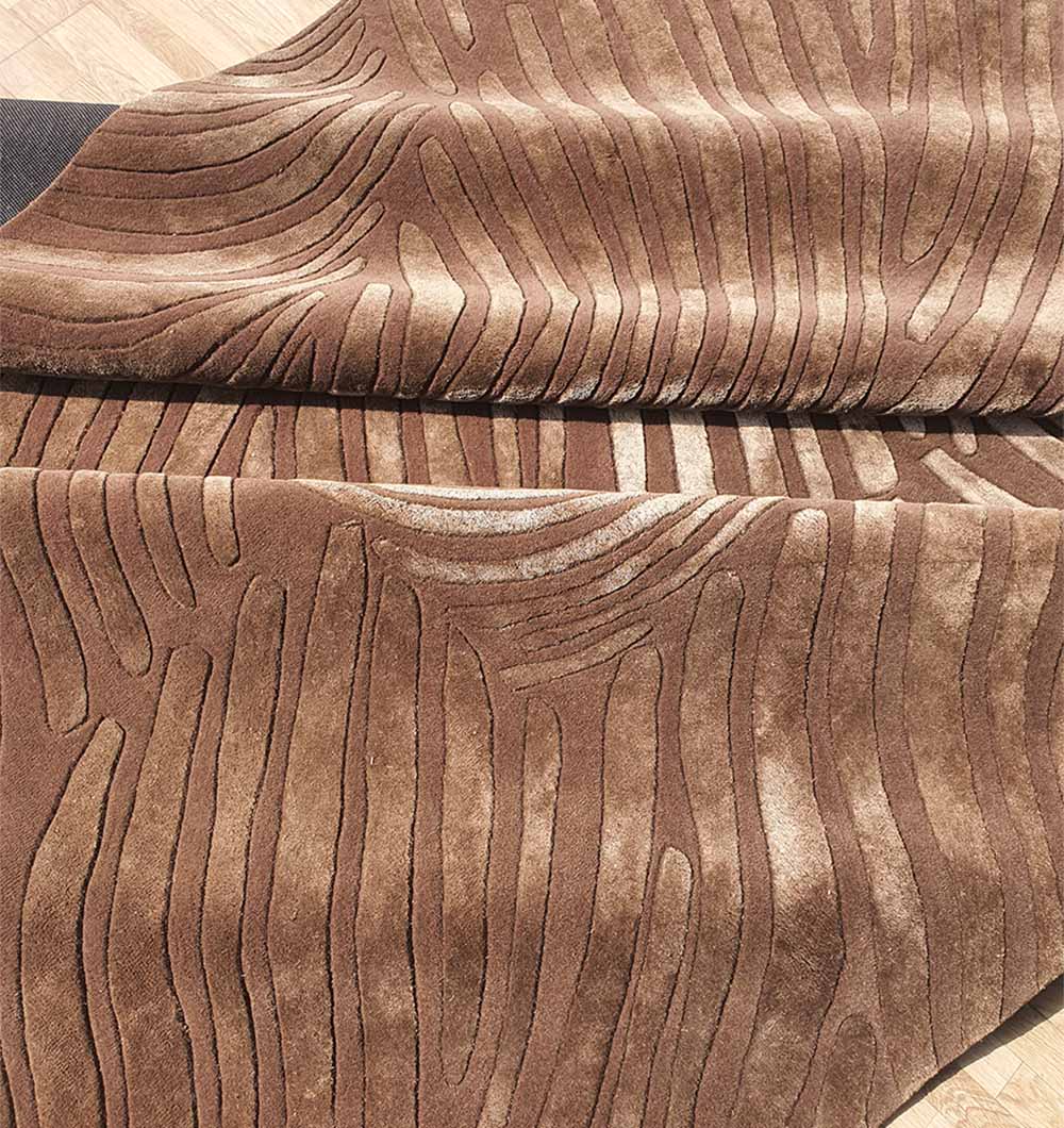 A close-up of the RAMSHA HOME Premium Quality Handmade Carpet HTWV-79 in chocolate brown, featuring a wavy wood grain-like embossed texture, partially folded and displayed on a wooden floor.