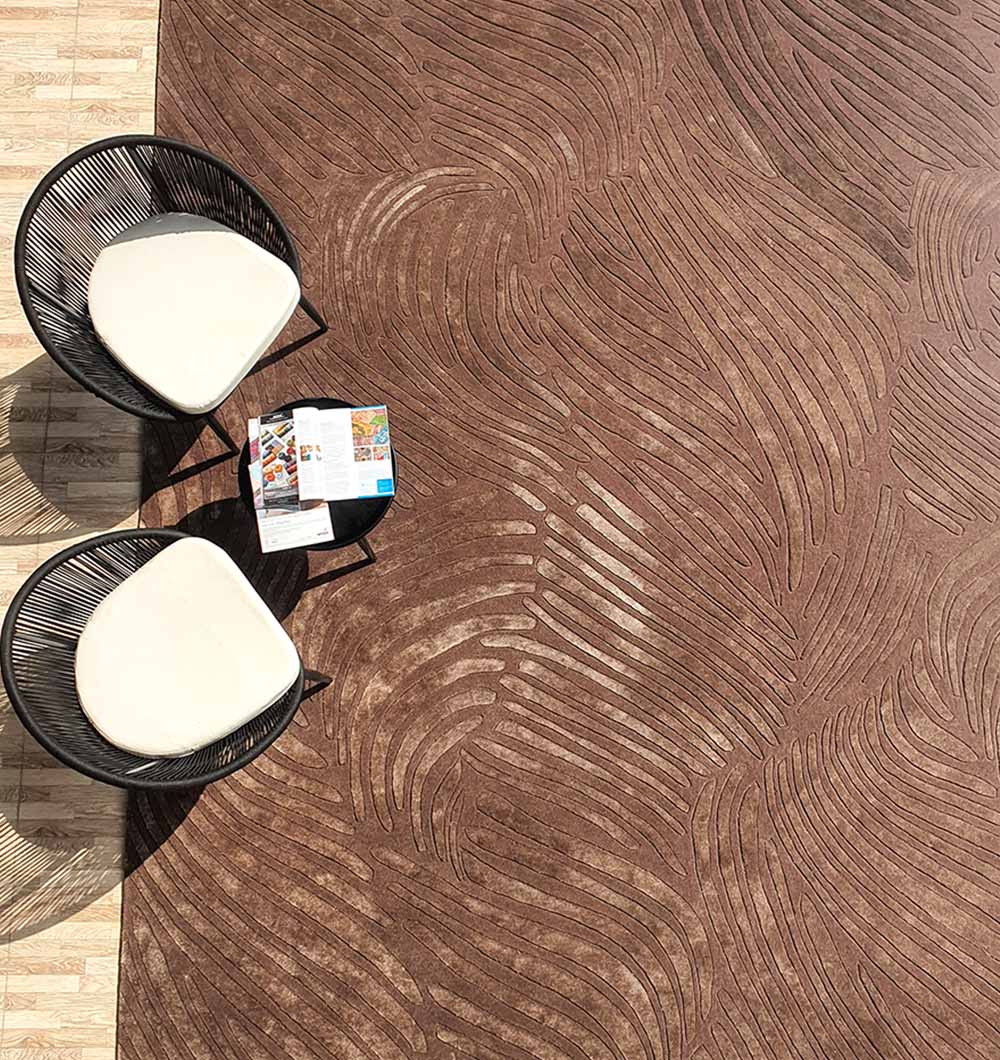 Two black-framed chairs with white cushions sit by a small round table holding an open magazine on the RAMSHA HOME Premium Quality Handmade Carpet HTWV-79, featuring chocolate brown abstract wavy patterns. A tiled floor is also visible.