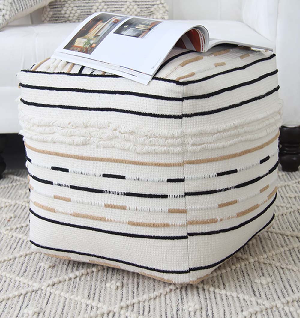 The RAMSHA HOME Cotton Boho Pouf PF-066, featuring square shape and beige, black, and white stripes, rests on a textured rug with an open magazine atop it. A white armchair appears partially in the background.