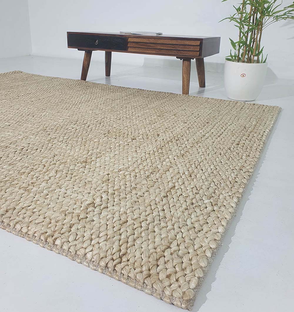 The RAMSHA HOME Bleach Color Woven Floor Rug For Home Decor SR-037 rests on a white floor, paired with a wooden table and a potted plant for a minimalistic, modern look.