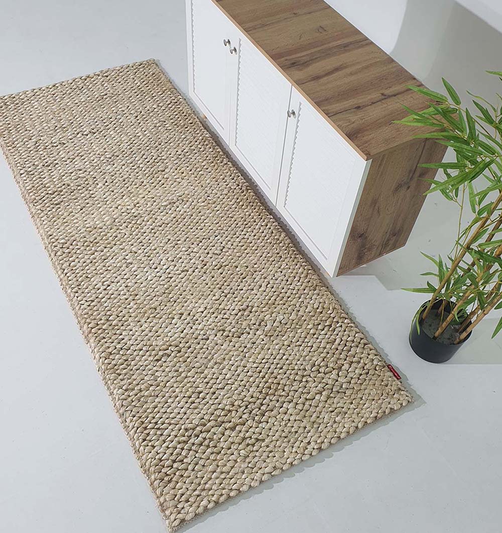 The RAMSHA HOME Bleach Color Woven Floor Rug For Home Decor SR-037 in beige jute rests on a white floor by a white cabinet with a wooden top, accented by a nearby potted bamboo plant.