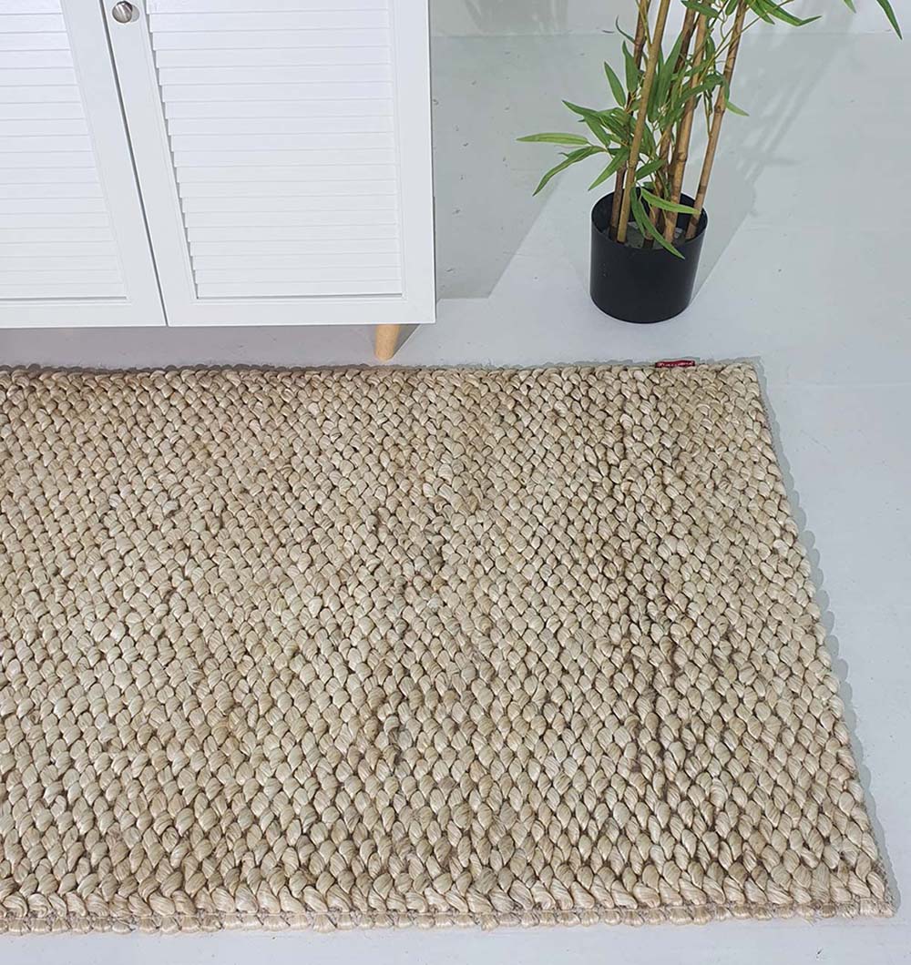 The RAMSHA HOME Bleach Color Woven Floor Rug For Home Decor SR-037, crafted from beige jute, is displayed on a white floor beside a cabinet with wooden legs and a potted green plant in a black container.