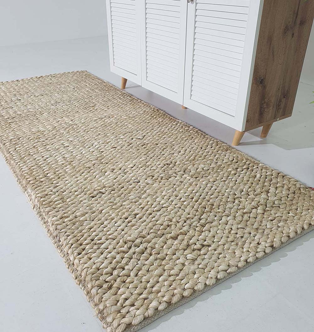 The RAMSHA HOME Bleach Color Woven Floor Rug For Home Decor SR-037, a rectangular handmade jute rug, is set on a white floor beside a wooden cabinet with white louvered doors and angled legs.