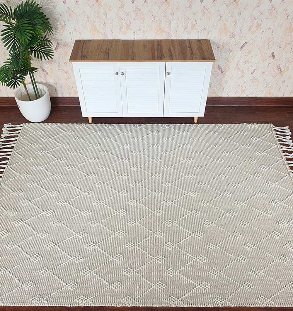 The RAMSHA HOME Designer Room Woven Rug Wool Natural Color SRN-005, featuring a beige geometric pattern and fringed edges, is laid on a wooden floor. Behind it, a white cabinet and potted plant stand against a textured cream wall.