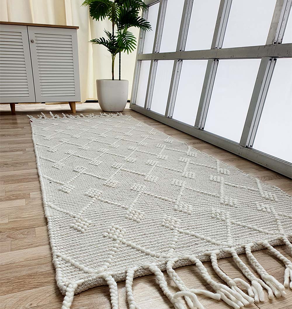 The RAMSHA HOME Designer Room Woven Rug Wool Natural Color SRN-005 with a geometric diamond pattern and tasseled edges lies on a wooden floor beside a white cabinet, potted plant, and large sunlit windows.