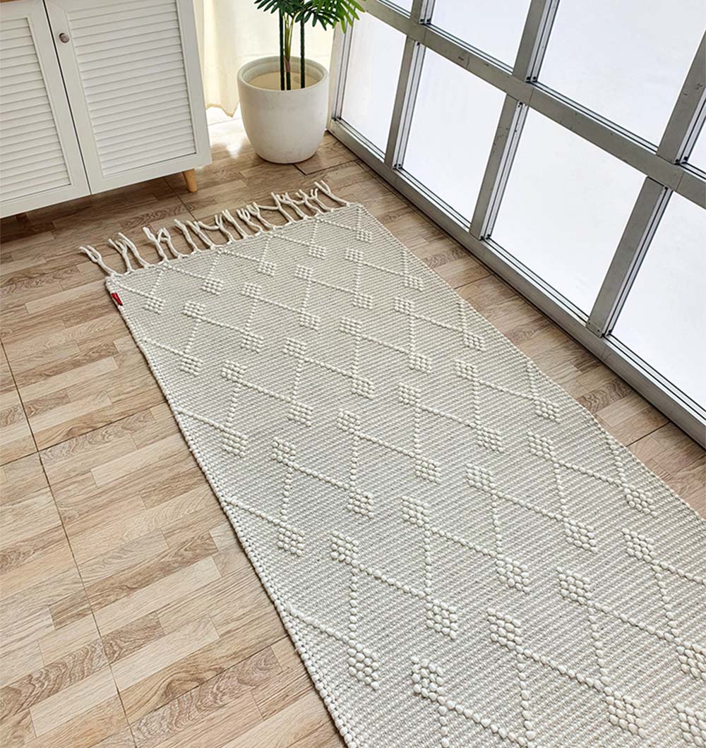 The RAMSHA HOME Designer Room Woven Rug Wool Natural Color SRN-005, featuring a textured geometric diamond pattern in cream, is placed on a light wood floor beside a white cabinet, potted plant, and sunlit windows.