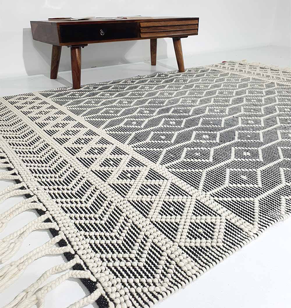 A close-up of the RAMSHA HOME Hand Woven Fringes Rug Natural & Black Color SRN-018, featuring textured geometric patterns and fringed edges, displayed on a white floor beside a wooden coffee table with angled legs.