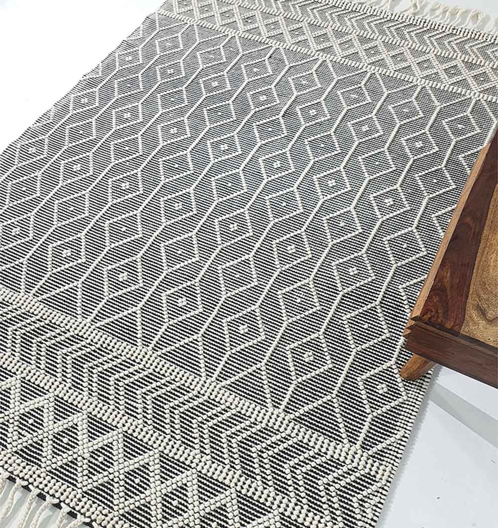 The RAMSHA HOME Hand Woven Fringes Rug (SRN-018) features a natural and black diamond pattern with fringed edges, shown with part of a wooden table on the right side.