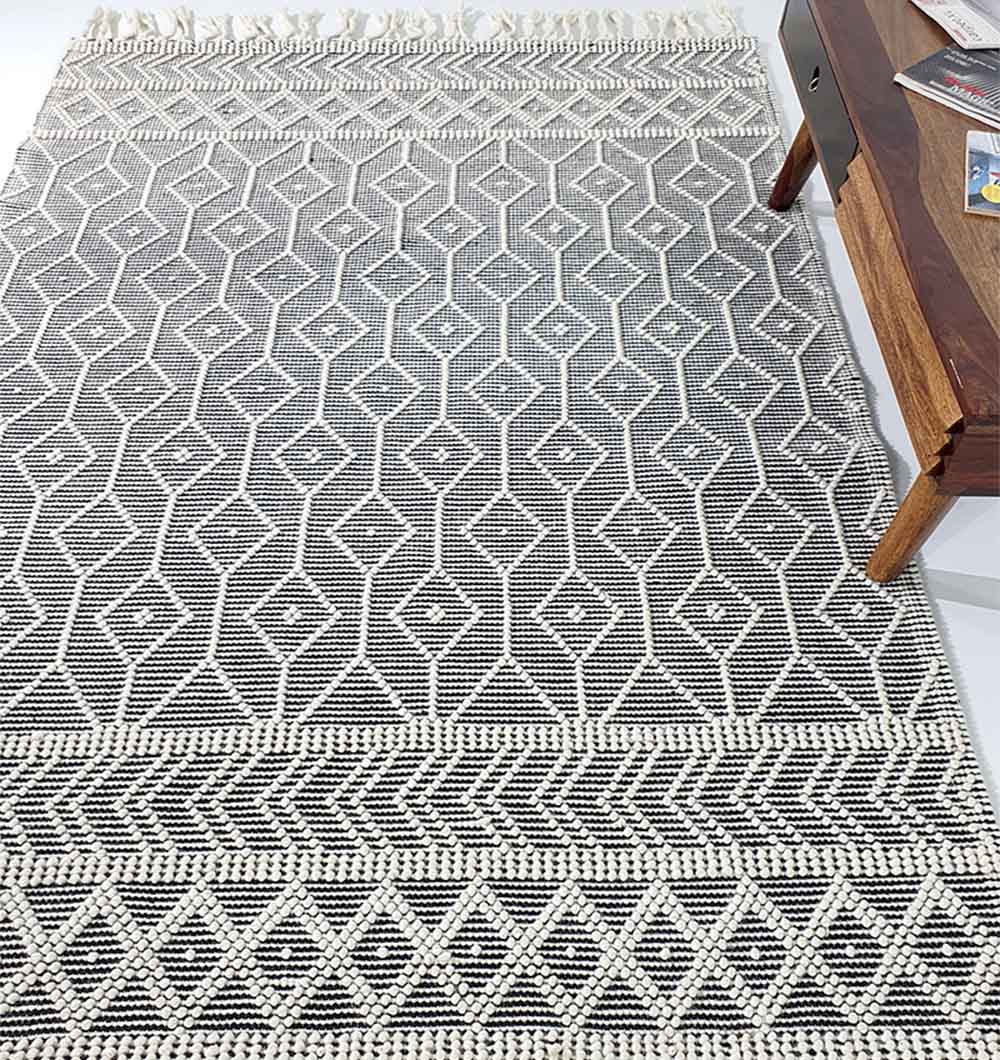 The Hand Woven Fringes Rug Natural & Black Color SRN-018 by RAMSHA HOME, featuring modern diamond and triangle patterns, lies on the floor beside a wooden table topped with magazines.