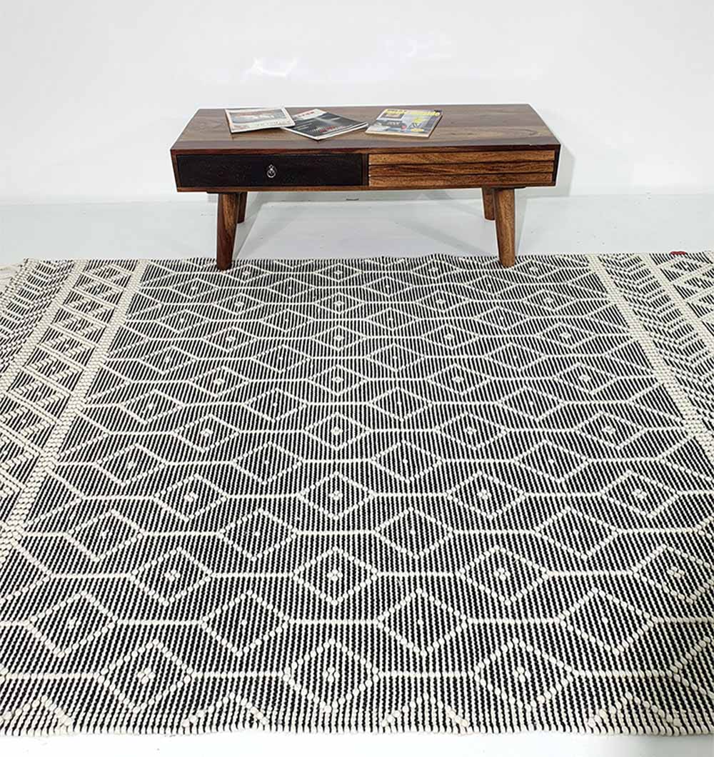 A RAMSHA HOME Hand Woven Fringes Rug in Natural & Black (SRN-018) with geometric diamond patterns lies under a mid-century modern wooden coffee table with magazines, framed by a plain white wall.