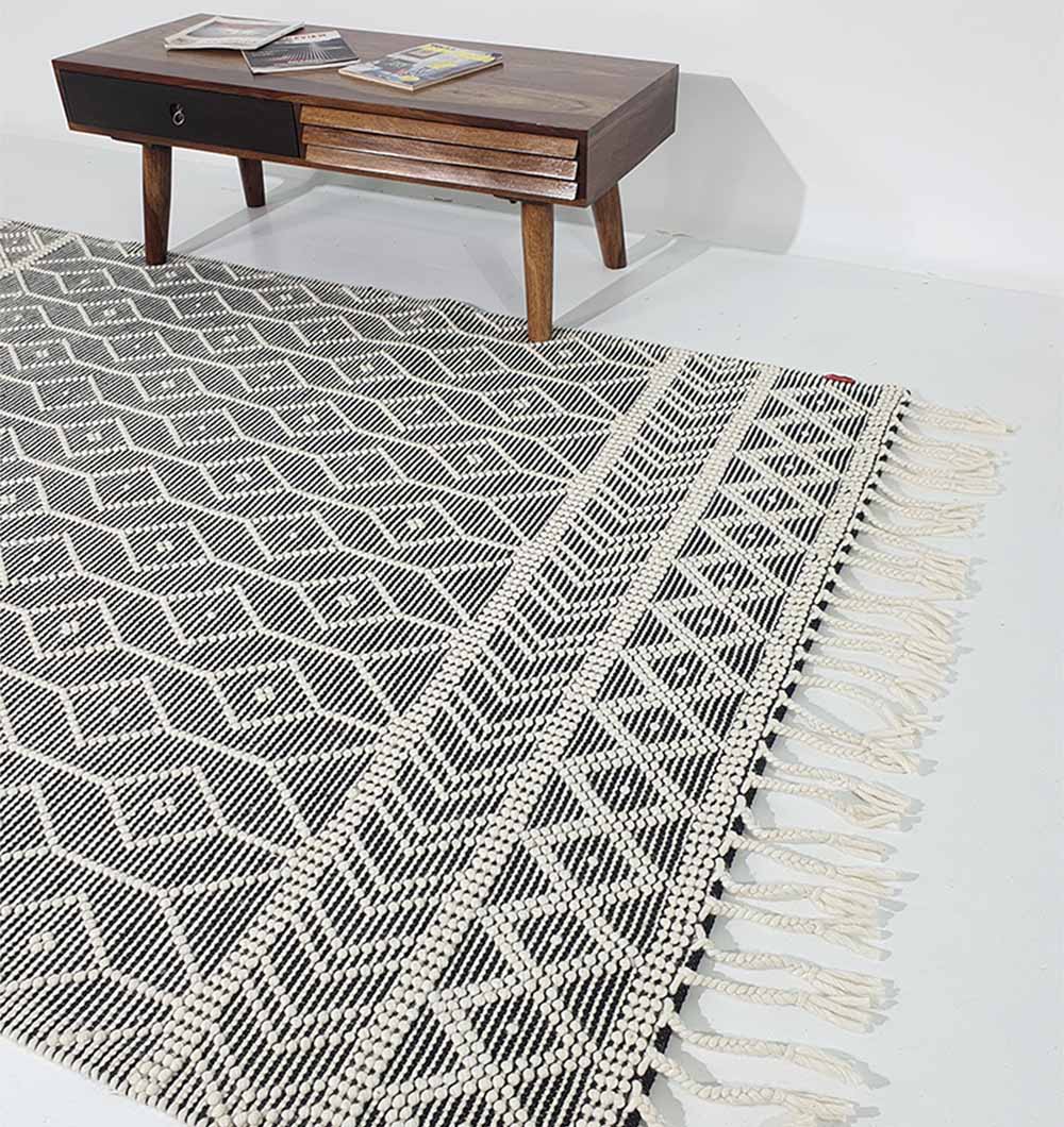 The RAMSHA HOME Hand Woven Fringes Rug Natural & Black Color SRN-018 is laid on the floor in front of a wooden coffee table displaying a magazine and a small item.