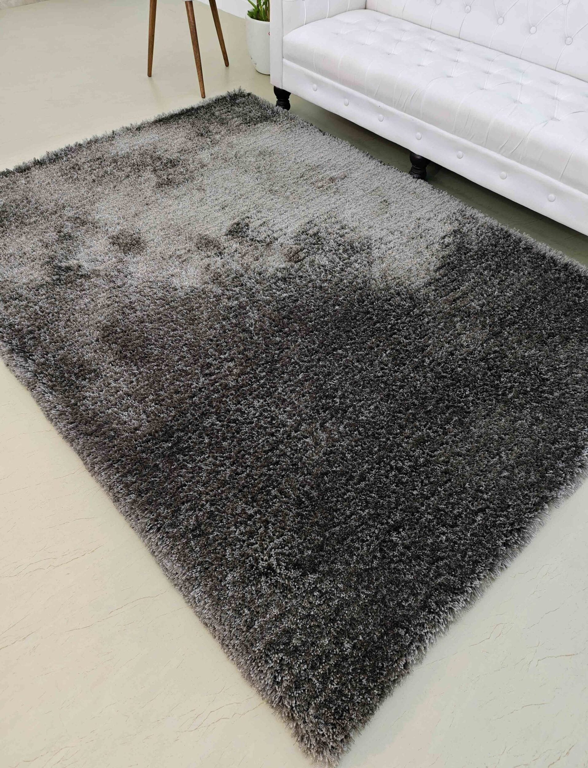 The RAMSHA HOME Grey Color Rectangle Shaped Shaggy Rug - SG 05 with a gradient effect is placed on a light floor beside a white tufted sofa and a small plant stand.