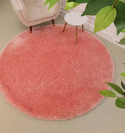 The RAMSHA HOME Round Shaped Pink Shaggy Rug - SG 01 rests on a light floor near a white armchair and small white side table with wooden legs, with green leaves from nearby plants partly visible in the foreground.
