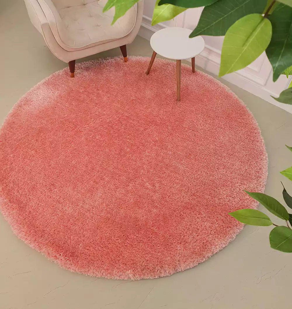 The RAMSHA HOME Round Shaped Pink Shaggy Rug - SG 01 rests on a light floor near a white armchair and small white side table with wooden legs, with green leaves from nearby plants partly visible in the foreground.