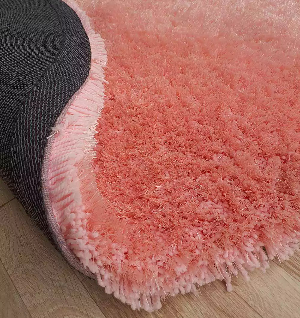 A close-up of the RAMSHA HOME Round Shaped Pink Shaggy Rug - SG 01 shows its plush coral pink pile and dark non-slip backing with a flipped corner, placed on a light wooden floor.
