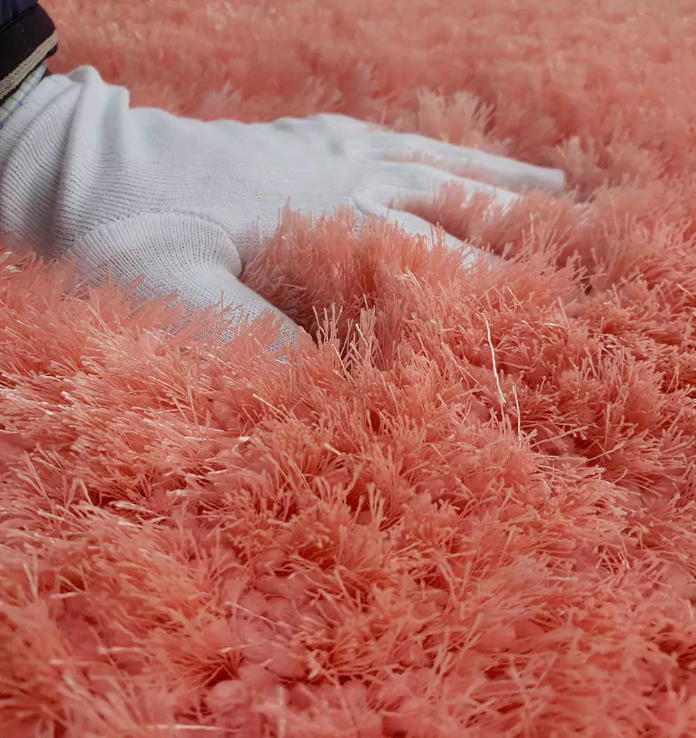 A gloved hand gently presses into the plush, long fibers of the RAMSHA HOME Round Shaped Pink Shaggy Rug - SG 01, highlighting its soft and fluffy coral pink texture.