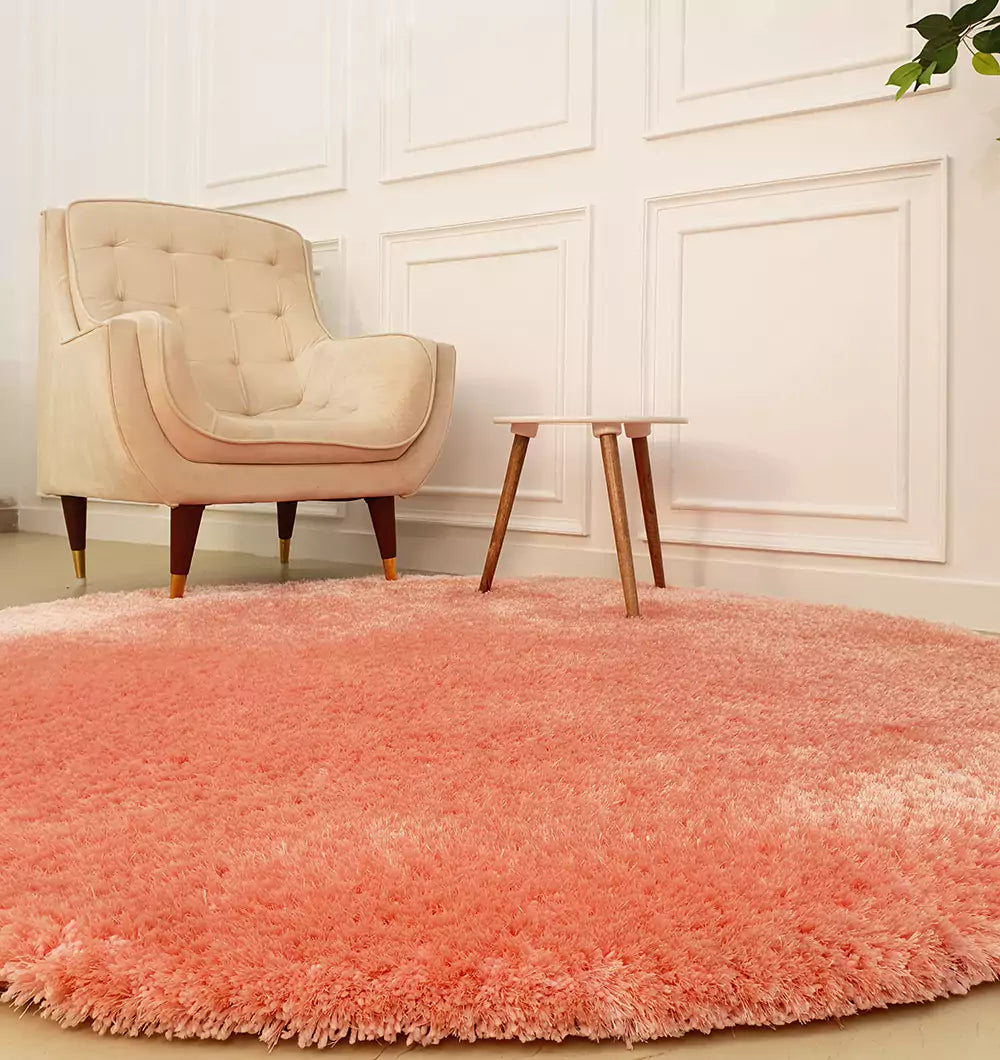 A modern living room includes the RAMSHA HOME Round Shaped Pink Shaggy Rug - SG 01, a beige tufted armchair with wooden legs, and a small wooden side table. White paneled walls form the backdrop with a potted plant partially visible.