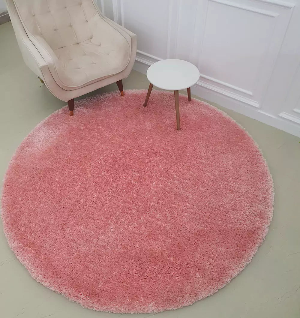 The RAMSHA HOME Round Shaped Pink Shaggy Rug - SG 01 adds a fluffy coral accent atop a light floor, paired with a beige tufted armchair and a white side table with wooden legs, all set before a crisp white paneled wall.