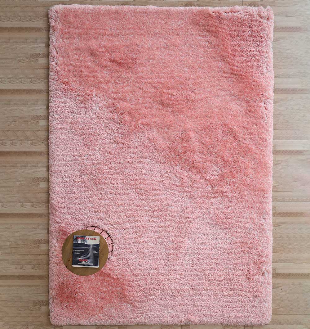 The RAMSHA HOME Pink Home Decor Shaggy Rug - SG 01, a soft rectangular blush rug, lies on a wooden floor. A circular cutout displays a folded magazine beneath, revealing the plush thickness and cozy pile of this area rug.