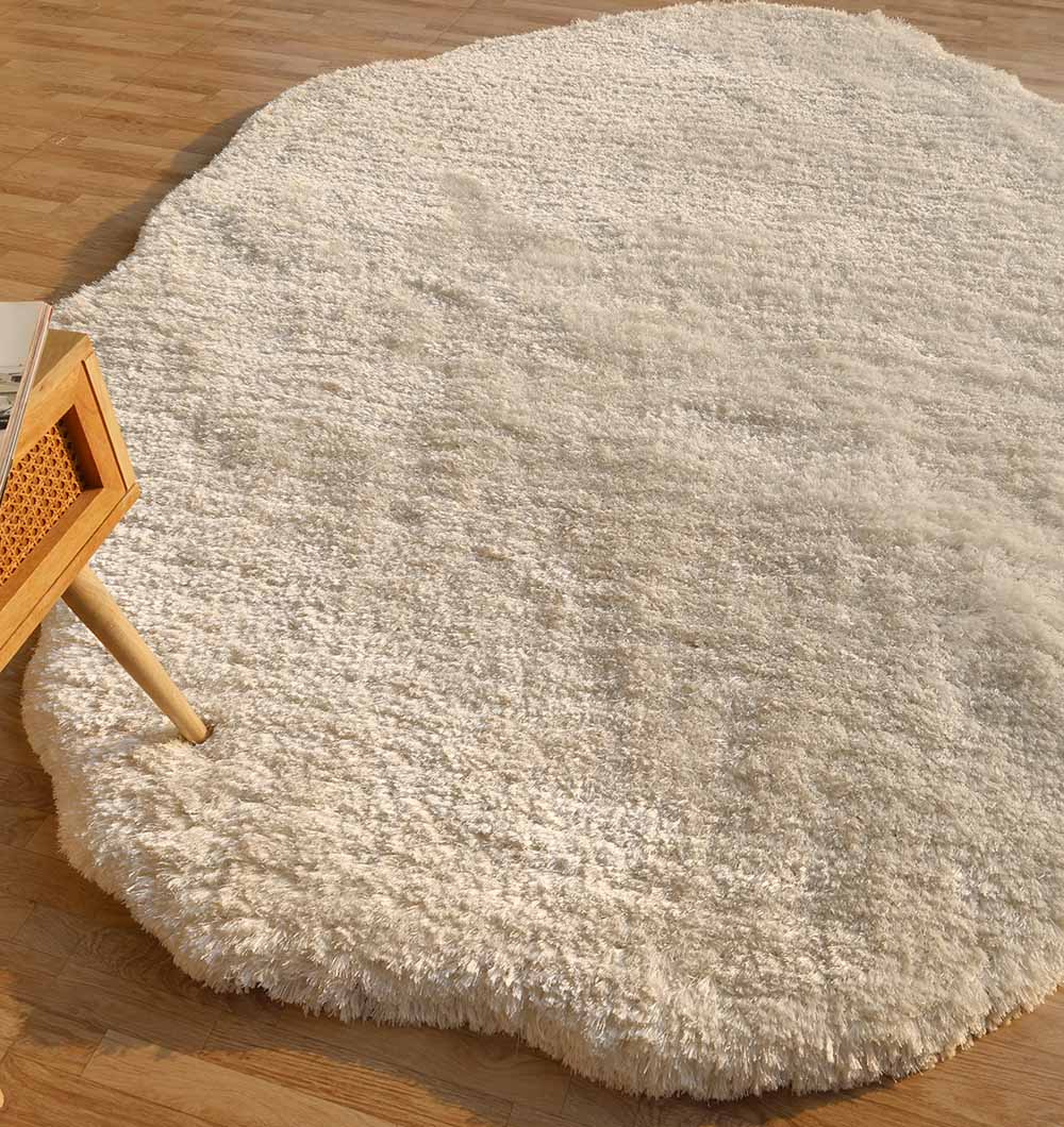 The RAMSHA HOME White Color Shape Shaggy Rug - SG 010 (200x300cm), with its plush pile and irregular shape, is placed on a wooden floor beside the corner of a light wood table.
