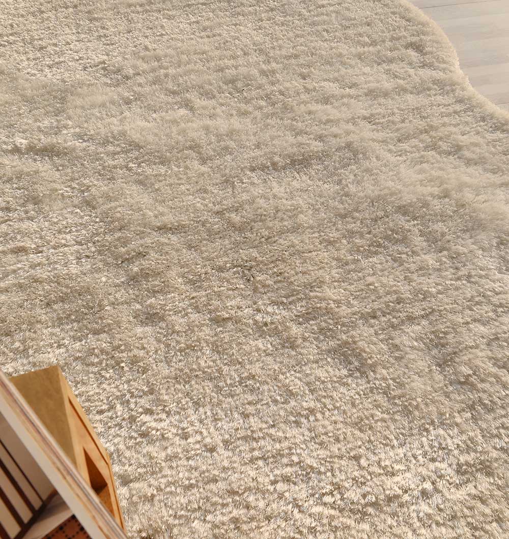 A close-up of the RAMSHA HOME White Color Shape Shaggy Rug - SG 010 (200x300cm) showing its plush pile on a light wood floor, with a wooden bookshelf corner in view.