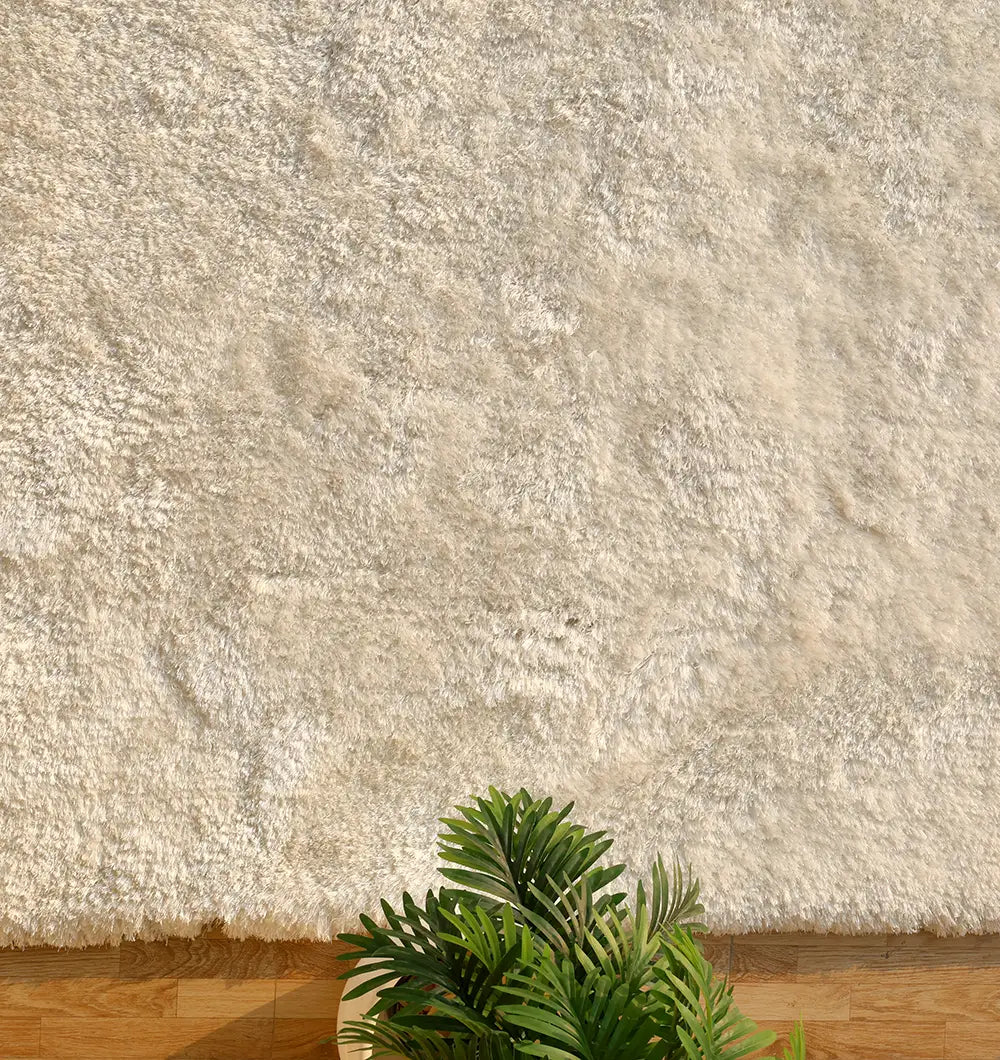 The RAMSHA HOME Plush White Shaggy Rug (SG-011) covers most of the image, while the corner of a potted plant with palm-like green leaves appears at the bottom edge on a wooden floor.