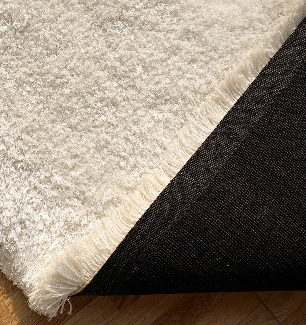 A close-up of the RAMSHA HOME Plush White Shaggy Rug for Modern Living Room Decor (SG-011), showing its fringed edges and black non-slip backing on a wooden floor.
