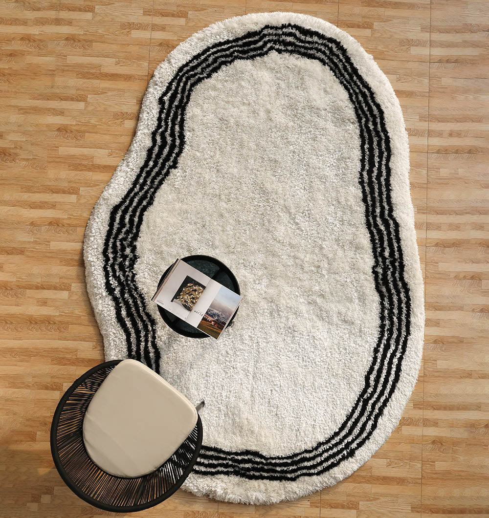 The RAMSHA HOME White & Black Shaggy Rug (SG-013) with black wavy lines is on a wooden floor, next to a round black side table with books and a modern chair featuring a black frame and light cushion.