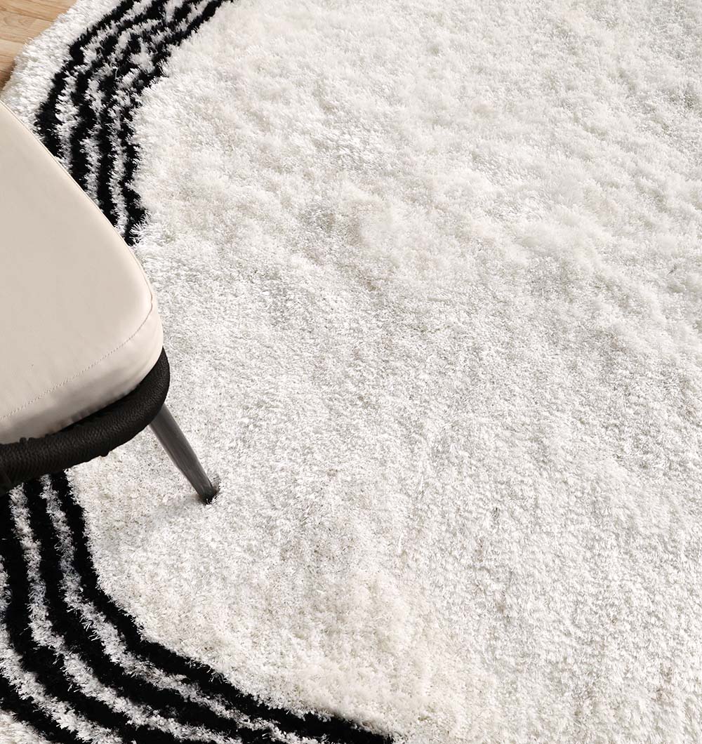 A close-up of the RAMSHA HOME White & Black Shaggy Rug for Modern Living Room Decor (SG-013) shows black curved lines on ivory. A beige cushioned chair with metal legs appears in the corner, highlighting the rug’s modern, stylish design.