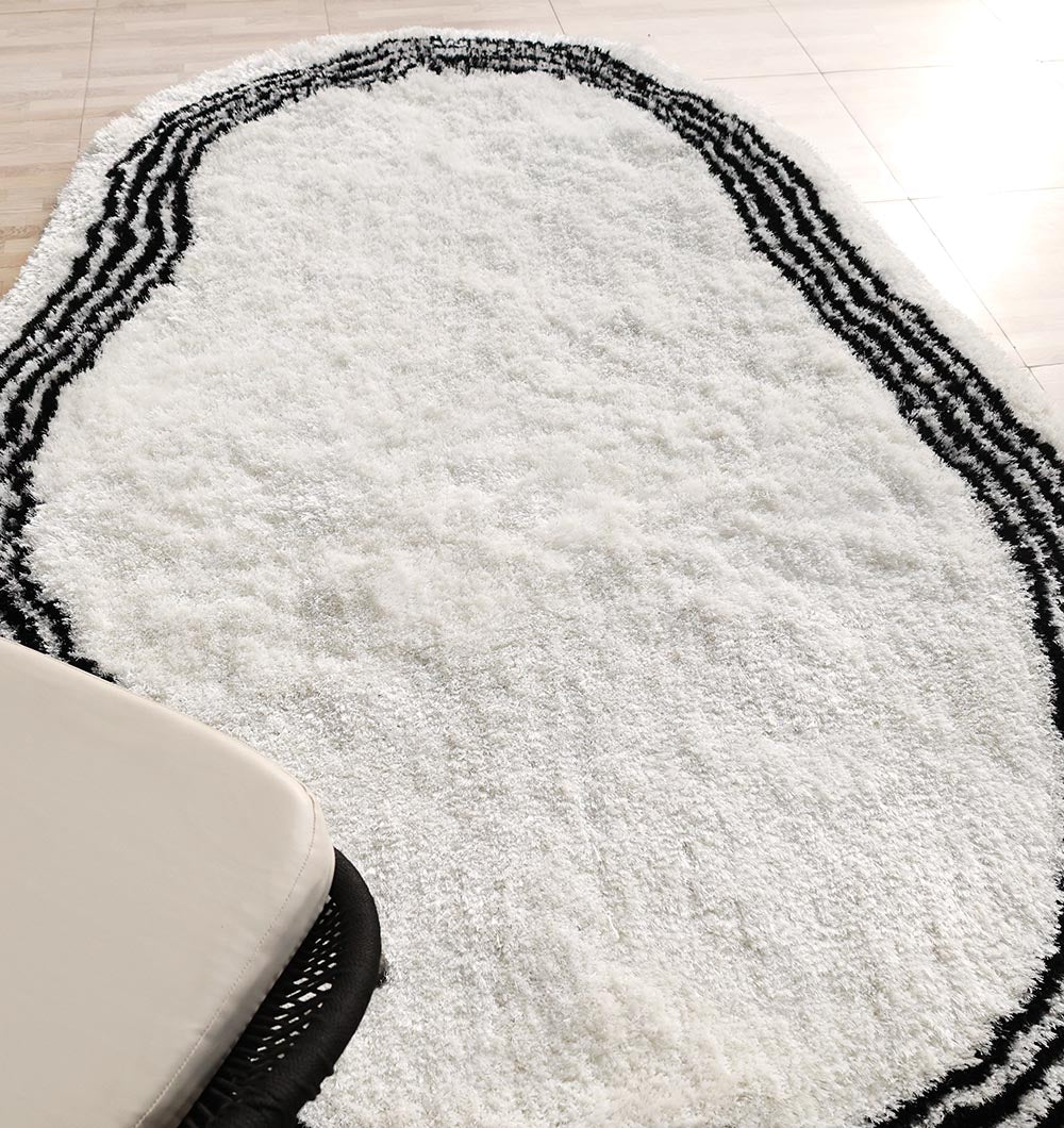 The RAMSHA HOME White & Black Shaggy Rug (SG-013), featuring an ivory base with irregular black wavy border lines, is styled on a light wooden floor next to a beige cushioned chair for modern living room decor.