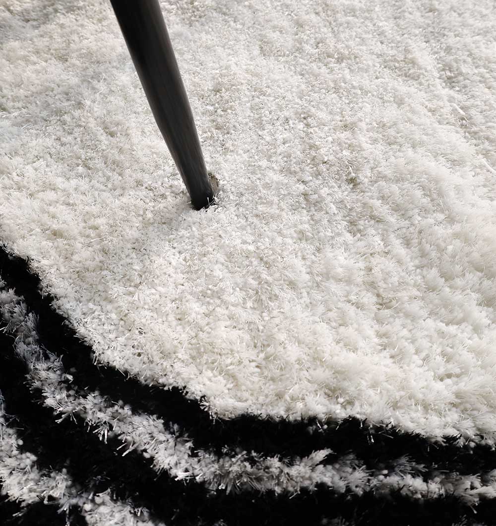 Close-up of the RAMSHA HOME White & Black Shaggy Rug for Modern Living Room Decor (SG-013), featuring plush ivory texture, a black border, and irregular shaggy edges with a table or chair leg resting on its soft surface.