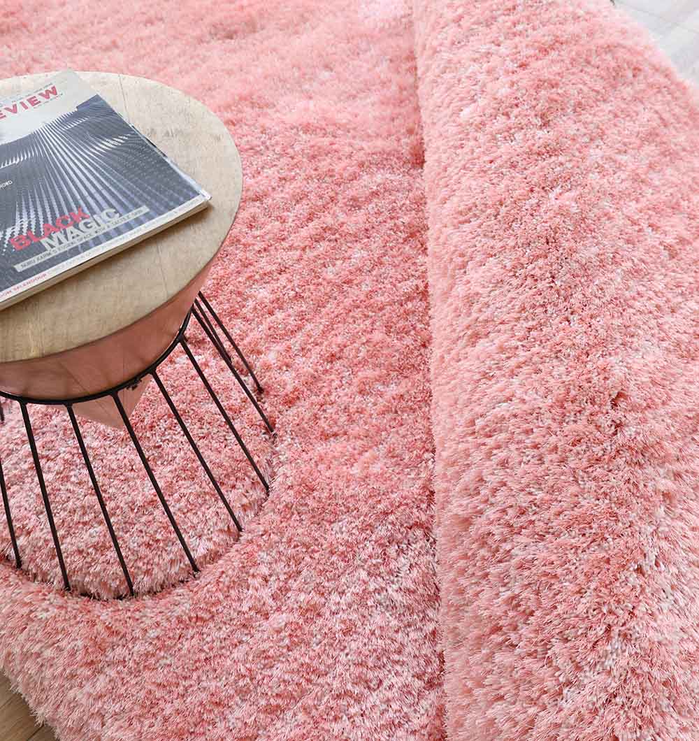 A close-up of the RAMSHA HOME Pink Home Decor Shaggy Rug - SG 01, featuring its soft, fluffy pink texture with a folded corner. A round wooden table with a wire base sits on the rug, holding a magazine.