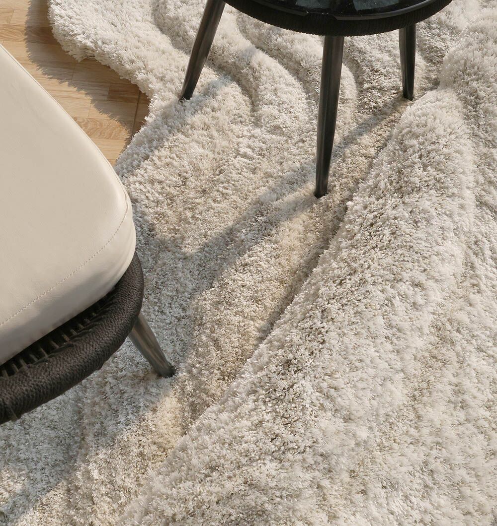 A close-up of the RAMSHA HOME Wavy Texture Shape Rug for Living Room - SG 016 (200 x 300 cm) shows its beige, irregular shape on a wooden floor, with chair and ottoman legs nearby as sunlight highlights its plush, textured surface.