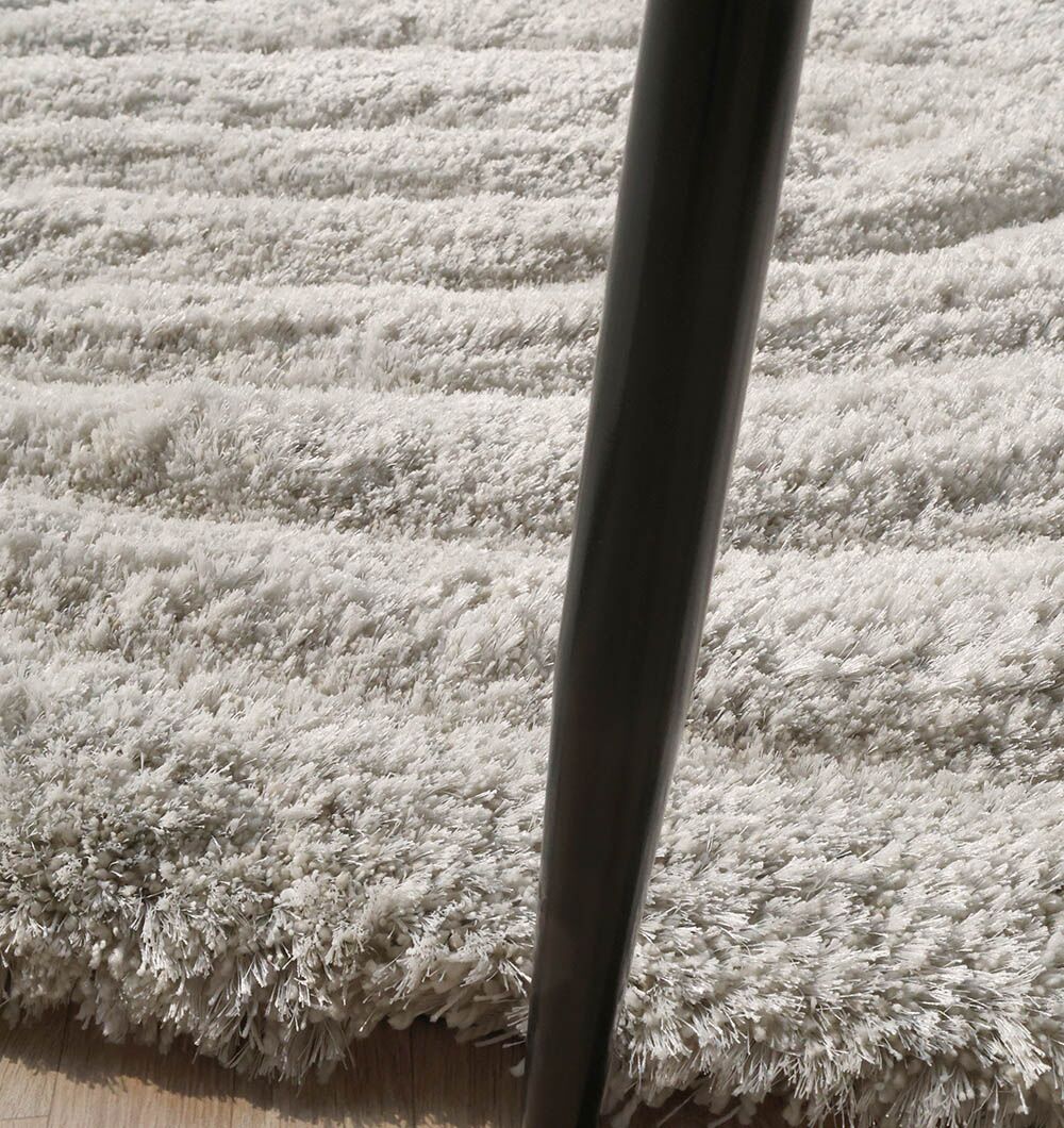A close-up of the RAMSHA HOME Wavy Texture Shape Rug for Living Room - SG 016 (200 x 300 cm) in light gray, with an irregular shape, is shown on a hardwood floor with a dark, slender furniture leg placed diagonally.