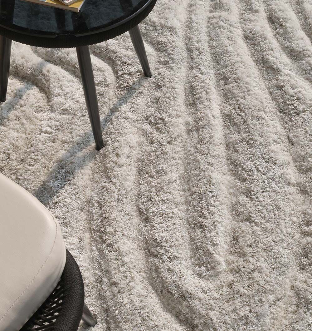 A close-up of the RAMSHA HOME Wavy Texture Shape Rug for Living Room - SG 016 (200 x 300 cm), paired with a round black table and beige chair. A book and eyeglasses on the table enhance the cozy vibe of this soft, irregular-shaped rug.