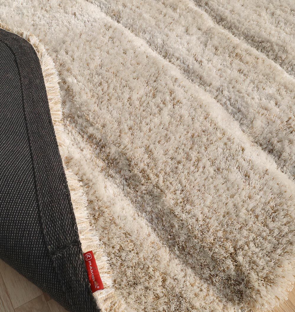 A close-up of the RAMSHA HOME Shaggy Shape Rug for Living Room - SG 017 (200x300 cm), featuring a plush beige pile, textured surface, black non-slip backing, and red edge label, displayed on a light wooden floor.