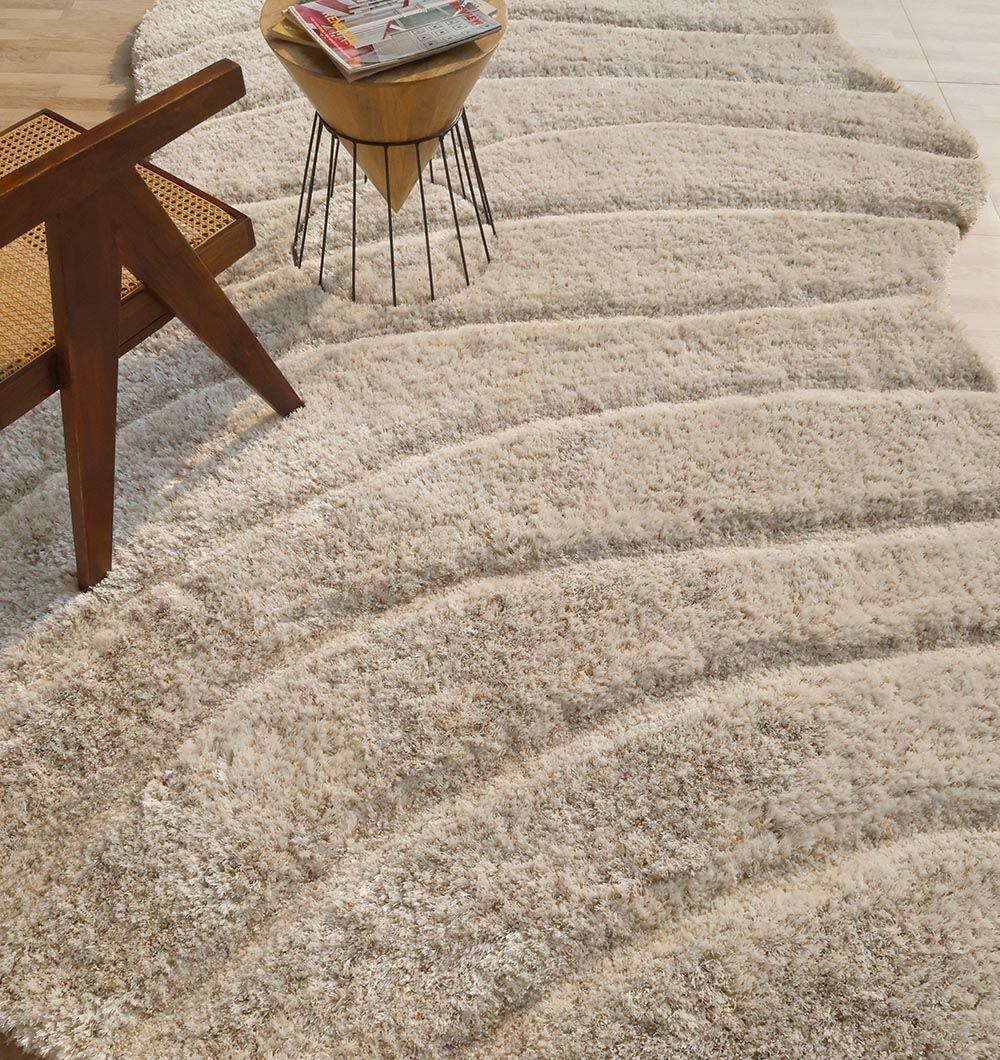The RAMSHA HOME Shaggy Shape Rug for Living Room - SG 017 (200x300 cm) in beige, featuring wavy raised patterns, is displayed on a light floor near a wooden chair with a woven seat and a side table with magazines.