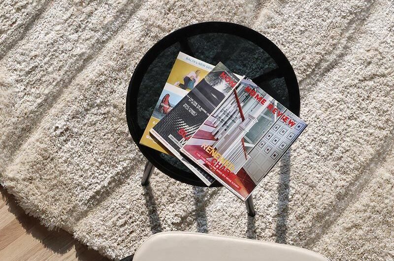 A round black glass coffee table with colorful magazines sits on a RAMSHA HOME Shaggy Shape Rug for Living Room - SG 018 (200X300 cm), as sunlight streams in. Part of a white chair appears in the lower foreground.