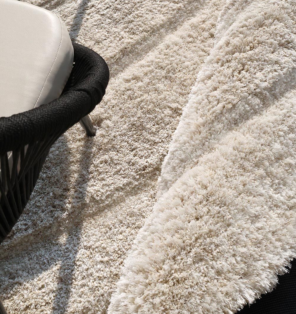 A close-up of the RAMSHA HOME Shaggy Shape Rug for Living Room - SG 018 (200x300 cm) in ivory-beige, with diagonal lines, partly covered by a black chair with a white cushion. Sunlight casts shadows across its soft, textured surface.