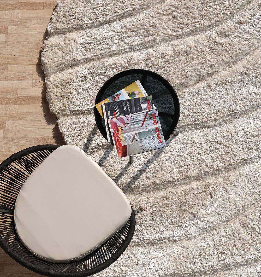 A round black side table with magazines sits on the RAMSHA HOME Shaggy Shape Rug for Living Room - SG 018 (200X300 cm), next to a chair with a cream cushion and black woven frame, on a light wood floor.